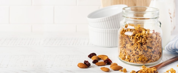 Granola owsiana z butelką mleka, orzechów i suszonych owoców, ceramiczne miseczki do przygotowania zdrowego śniadania na jasnym kuchennym stole. Skandynawski biały styl. Selektywna ostrość.