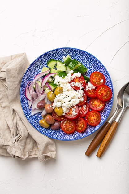 Grecka sałatka śródziemnomorska z pomidorami, serem feta, ogórkiem, całymi oliwkami i czerwoną cebulą w ceramicznym talerzu na białym kamiennym tle z góry, tradycyjna przystawka z Grecji