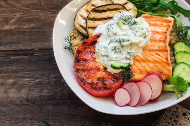 Grillowany łosoś, bakłażany i pomidory z quinoa i sosem tzatziki na rustykalnym drewnianym stole. Zdrowa kolacja.