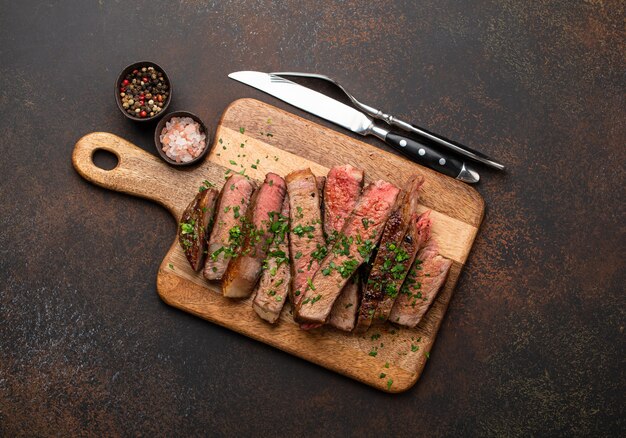 Grillowany lub smażony stek z doskonałego marmurkowego mięsa Ribeye pokrojony w plastry i podany na drewnianej desce do krojenia ze sztućcami. Widok z góry soczystego gotowanego steku wołowego pokrojonego w plastry na rustykalnym brązowym betonowym tle