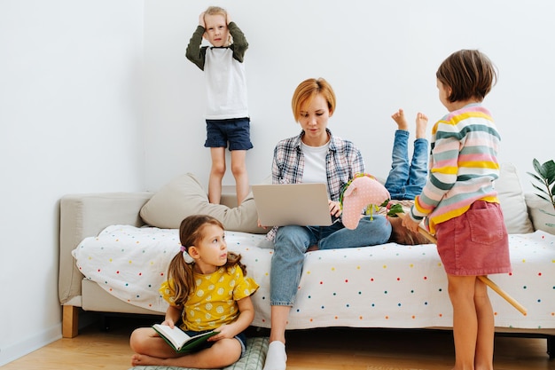 Grono dzieci bawi się wokół swojej mamy siedzącej na kanapie, pracującej na swoim laptopie