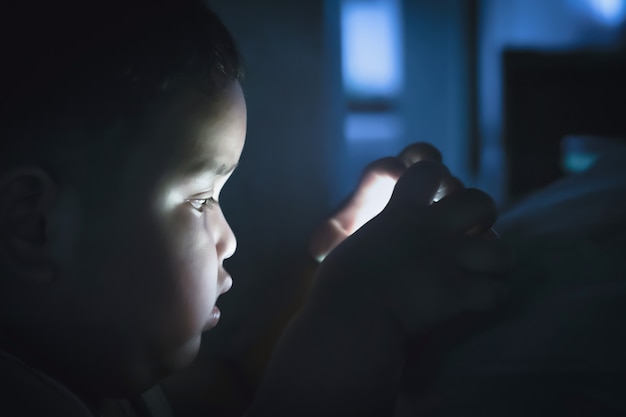 Gruba chłopiec bawić się smartphone w sypialni przy nighttime na ciemnym tle. Długotrwała gra telefoniczna negatywnie wpływa na wzrok i zdrowie małych dzieci.