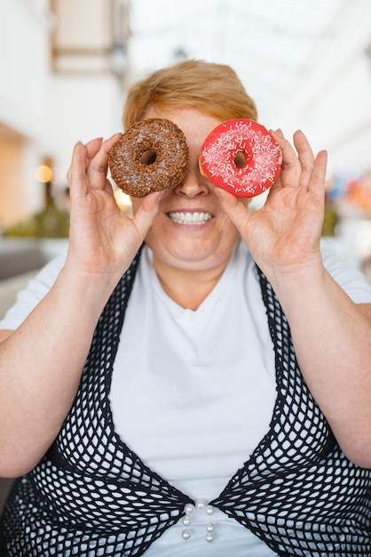 Zdjęcie gruba kobieta trzyma pączki zamiast oczu w restauracji w centrum handlowym, niezdrowa żywność. kobieta z nadwagą przy stole ze śmieciowym obiadem, problem z otyłością