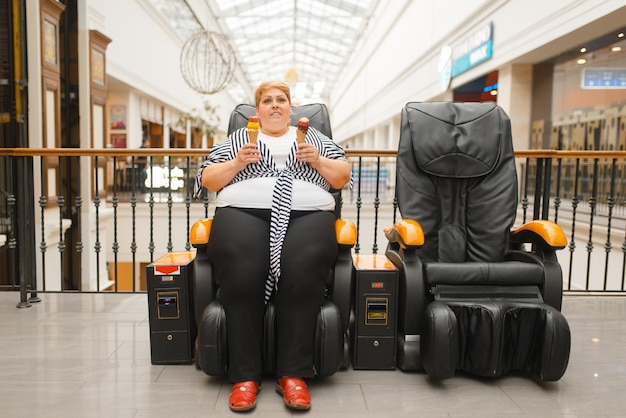 Gruba Kobieta Z Lodami Siedzi Na Fotelu Do Masażu W Centrum Handlowym. Kobieta Z Nadwagą Pozuje W Skórzanym Fotelu W Centrum Handlowym, Problem Z Otyłością