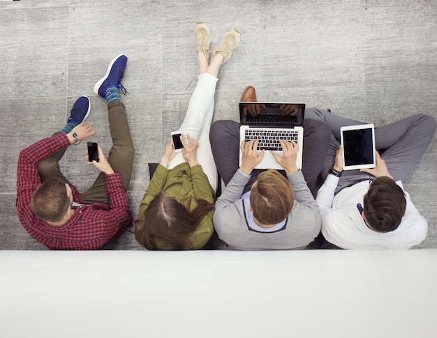 Grupa atrakcyjnych młodych ludzi siedzących na podłodze za pomocą laptopa, komputera typu Tablet, smartfonów, uśmiechając się.