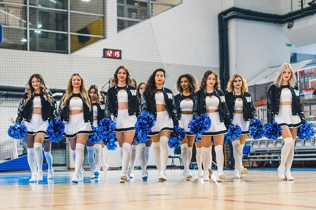 Grupa cheerleaderek w mundurach idących w kierunku kamery, trzymając niebieskie pompony