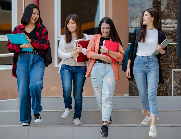 Grupa czterech młodych atrakcyjnych azjatyckich dziewcząt studentów idących razem po schodach w kampusie uniwersyteckim, rozmawiających i śmiejących się z radości. Koncepcja edukacji i życia studentów.