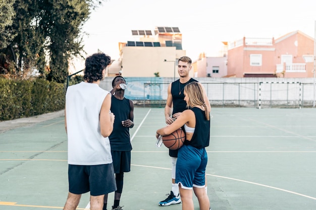 Grupa czterech wieloetnicznych przyjaciół stojących z piłką do koszykówki na boisku zewnętrznym