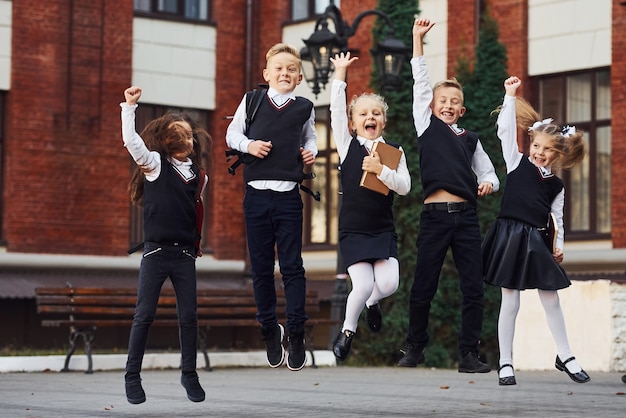 Grupa dzieci w mundurkach szkolnych, skoki i wspólna zabawa na świeżym powietrzu w pobliżu budynku edukacji.