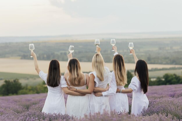 Grupa kobiet w białych sukienkach trzyma kieliszki wina na lawendowym polu