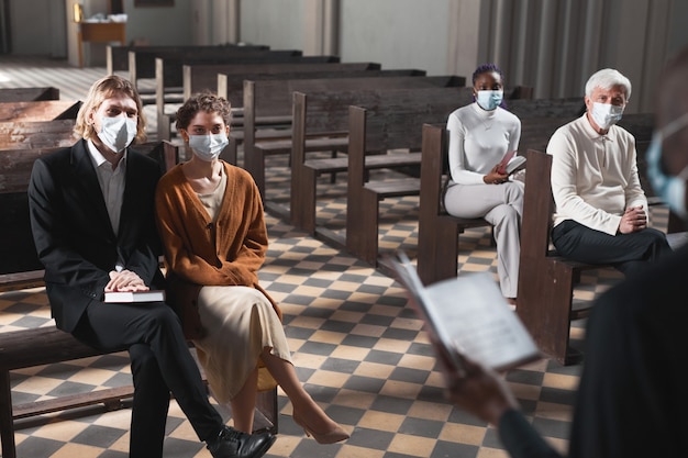 Grupa ludzi w maskach ochronnych siedząca na ławce i słuchająca mszy w kościele podczas pandemii