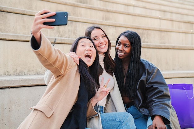 Grupa młodej kobiety, która dobrze się bawi, robiąc selfie ze smartfonem Trzy wielorasowe szczęśliwe dziewczyny