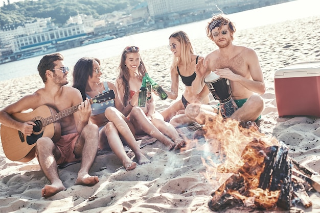 Grupa młodych koleżanek siedzących na plaży na leżakach i grających na gitarze, ciesząc się o zachodzie słońca