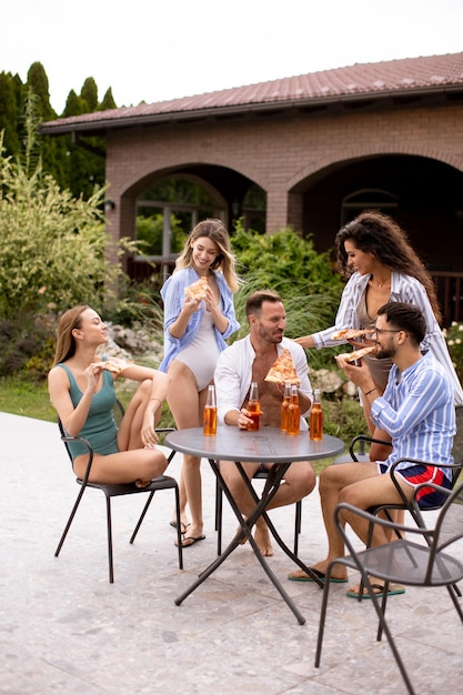 Grupa młodych ludzi dopingujących cydrem i jedzących pizzę przy basenie w ogrodzie