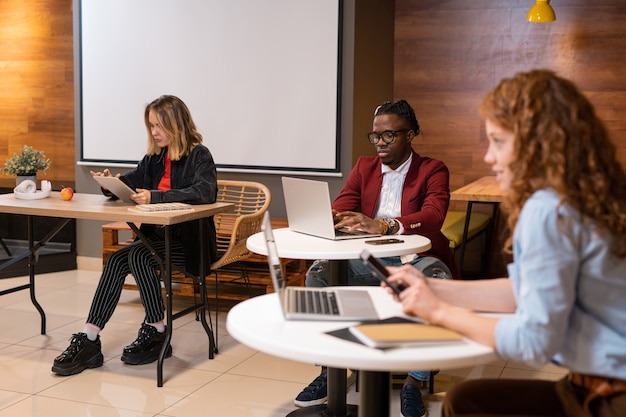 Grupa młodych międzykulturowych studentów college'u lub uniwersytetu siedząca przy stolikach w kawiarni i korzystająca z gadżetów podczas odrabiania lekcji