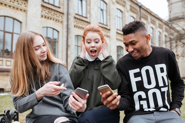 Grupa młodych przyjaznych wieloetnicznych studentów patrzących na telefon komórkowy siedząc na zewnątrz