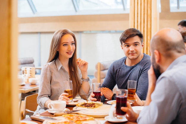 Grupa młodych wesołych przyjaciół siedzi w kawiarni rozmawiając i jedząc pizzę. Obiad w pizzerii.