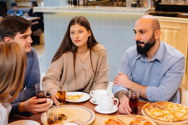 Grupa młodych wesołych przyjaciół siedzi w kawiarni rozmawiając i jedząc pizzę. Obiad w pizzerii.