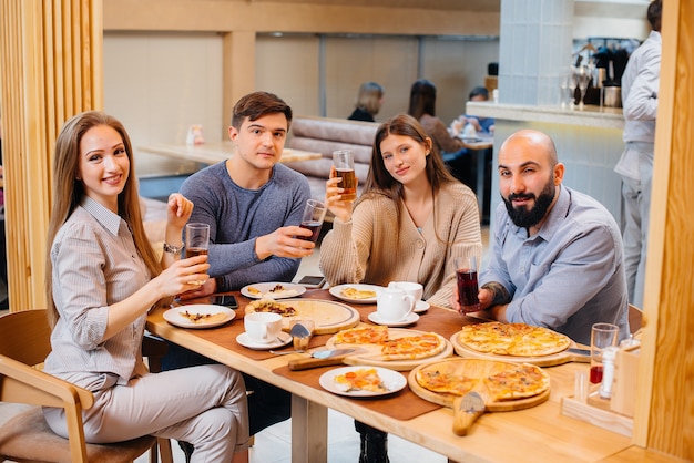 Grupa młodych wesołych przyjaciół siedzi w kawiarni rozmawiając i jedząc pizzę. Obiad w pizzerii.