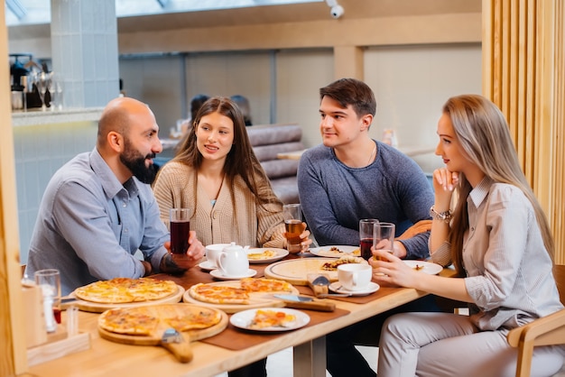 Grupa młodych wesołych przyjaciół siedzi w kawiarni rozmawiając i jedząc pizzę