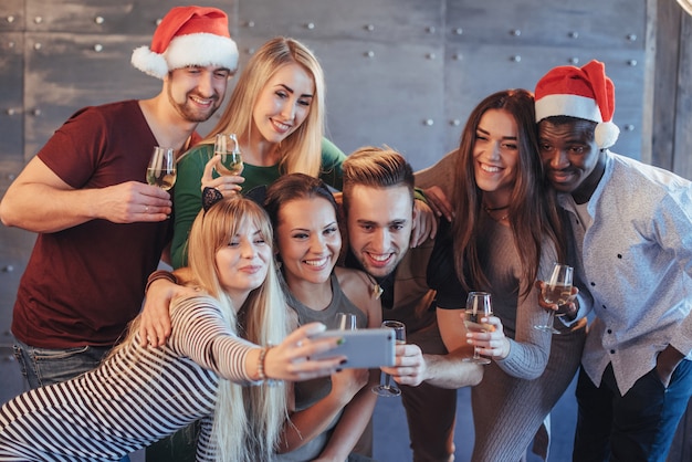 Grupa pięknych młodych ludzi robi selfie na imprezie noworocznej, najlepsi przyjaciele dziewczyny i chłopcy bawią się razem, stwarzając emocjonalny styl życia. Kapelusze Mikołaje i kieliszki do szampana w rękach