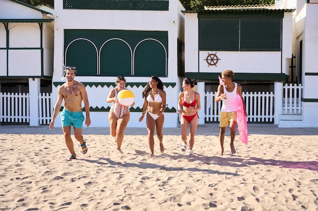Grupa podekscytowanych różnych przyjaciół biegnących wzdłuż piasku na miejskiej plaży w strojach kąpielowych