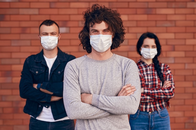 Grupa poważnych mężczyzn i kobiet ze skrzyżowanymi rękami w maskach medycznych i patrzących w kamerę podczas epidemii koronawirusa na mur z cegły