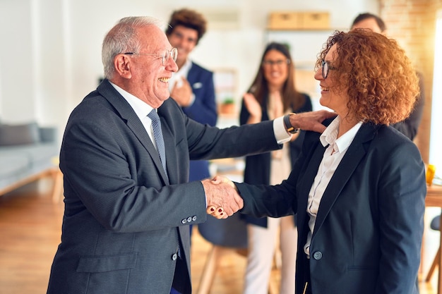 Grupa pracowników biznesowych uśmiecha się szczęśliwy i pewny siebie. Dwóch z nich stoi z uśmiechem na twarzy, ściskając dłonie za porozumieniem i partnerami klaszczącymi w biurze.