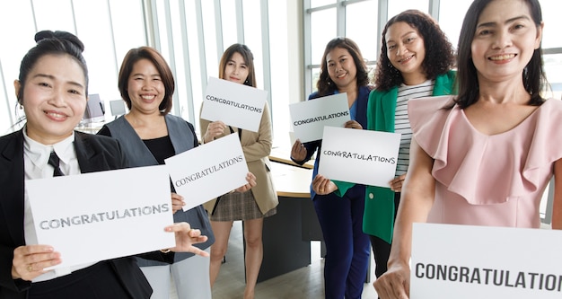 Grupa przedsiębiorców w ubrania biznesowe trzymając papierowy baner z gratulacjami słownymi i pokaż do kamery. Koncepcja powitania i uznania za sukces w pracy lub pracy w firmie.