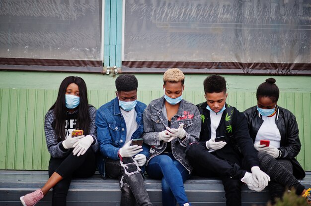 Grupa przyjaciół afrykańskich nastolatków siedzących z telefonami w maskach medycznych chroni przed infekcjami i chorobami kwarantanny wirusa koronawirusa.