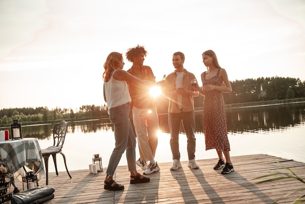 Grupa przyjaciół bawiąca się na pikniku nad jeziorem lub rzeką, pijąca wino, piwo, cydr, brzęk kieliszków podczas wznoszenia toastu. Uśmiechnięci młodzi ludzie bawią się na świeżym powietrzu podczas zachodu słońca na wsi.