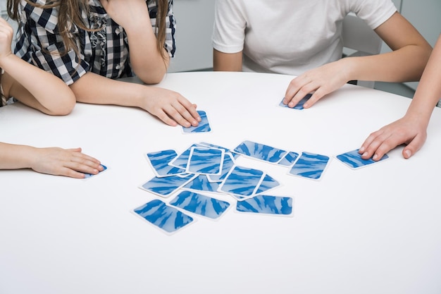 Grupa przyjaciół gra w karty siedząc przy białym stole Dzieci biorą ze stosu odwrócone papierowe niebieskie karty