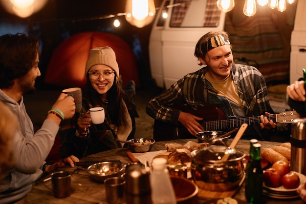 Grupa przyjaciół siedzi przy stole i gra na gitarze wieczorem podczas pikniku na świeżym powietrzu