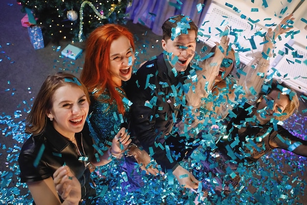 Zdjęcie grupa przyjaciół świętuje nowy rok konfetti w pobliżu choinki. ludzie śmieją się i radują.