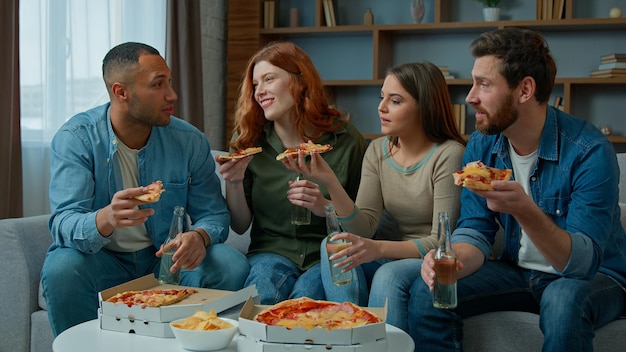 Grupa różnorodnych przyjaciół jedzących pizzę, rozmawiających szczęśliwie i pijących piwo świętujących imprezę