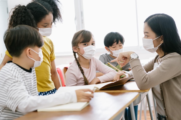 Grupa różnorodnych uczniów szkoły podstawowej i azjatyckiej nauczycielki noszącej maskę medyczną w szkole podstawowej Nauczyciel pomagający uczniom w klasieSzkoła ponownie otwarta po kwarantannie i blokadzie covid19
