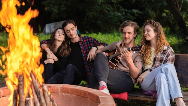 Grupa szczęśliwych młodych przyjaciół przy ognisku w glampingu. Dwóch mężczyzn i kobiet. Grać na gitarze