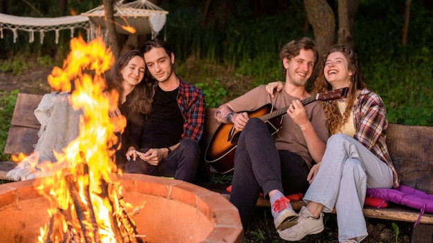 Grupa szczęśliwych młodych przyjaciół przy ognisku w glampingu. Dwóch mężczyzn i kobiet. Grać na gitarze