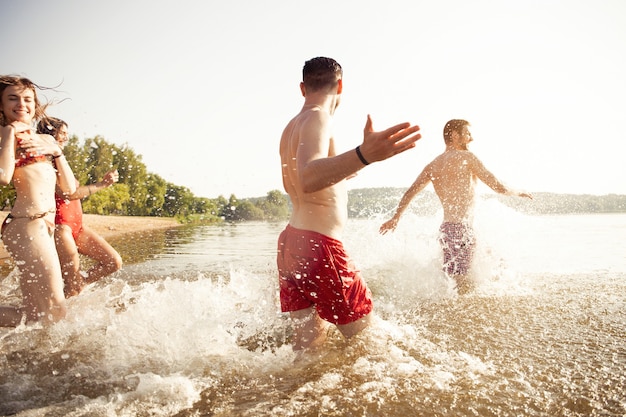Grupa szczęśliwych przyjaciół biegnących do wody - aktywni ludzie bawią się na plaży na wakacjach - turyści chodzą popływać
