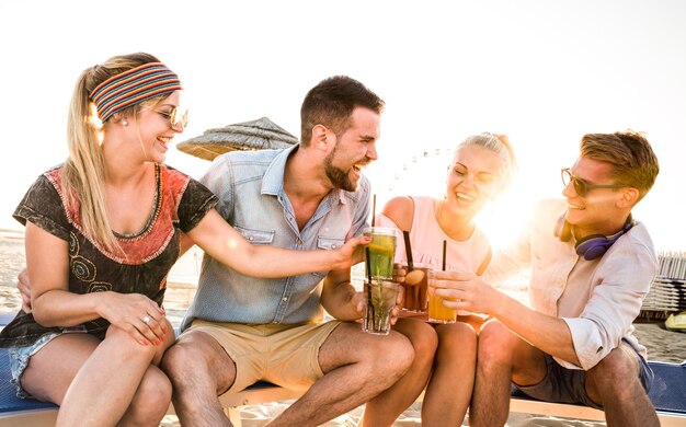 Grupa szczęśliwych przyjaciół milenijnych bawiących się na imprezie na plaży, popijając koktajl o zachodzie słońca