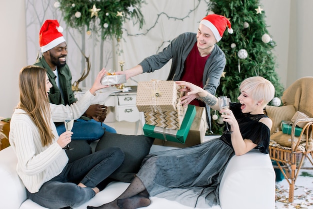 Grupa szczęśliwych przyjaciół siedzi na białej kanapie i picia szampana, trzymając prezenty świąteczne