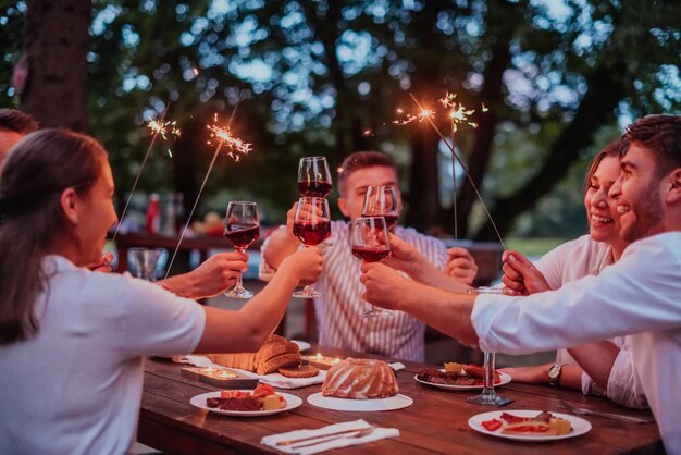 Grupa szczęśliwych przyjaciół świętujących wakacje za pomocą zraszaczy i pijących czerwone wino podczas piknikowej francuskiej kolacji na świeżym powietrzu w pobliżu rzeki w piękny letni wieczór w przyrodzie.