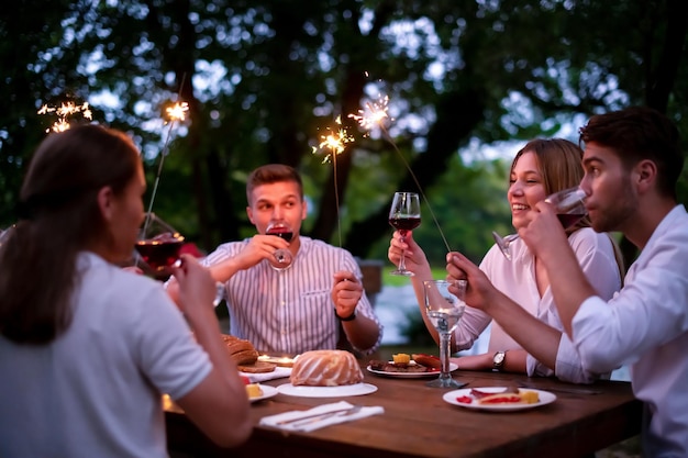 grupa szczęśliwych przyjaciół świętujących wakacje za pomocą zraszaczy i pijących czerwone wino podczas pikniku francuska kolacja na świeżym powietrzu w pobliżu rzeki w piękny letni wieczór w przyrodzie