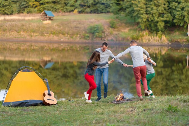 Grupa szczęśliwych przyjaciół z gitarą, zabawy na świeżym powietrzu, tańca i skakania w pobliżu jeziora w parku Piękne niebo. Zabawa na kempingu