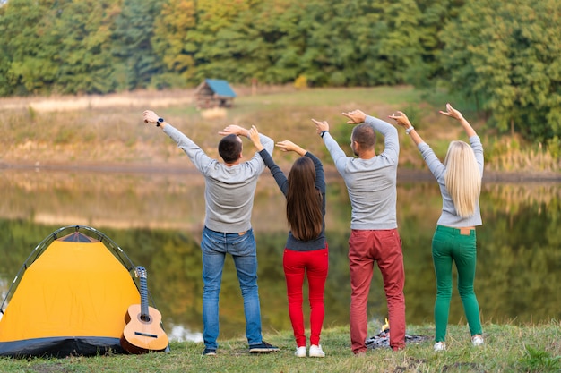 Grupa szczęśliwych przyjaciół z gitarą, zabawy na świeżym powietrzu, taniec trzymać ręce w pobliżu jeziora w tle parku Piękne niebo. Zabawa na kempingu