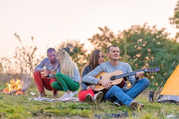 Grupa szczęśliwych przyjaciół z gitarą, zabawy na świeżym powietrzu, w pobliżu ogniska i namiotu turystycznego. Zabawa na kempingu szczęśliwa rodzina