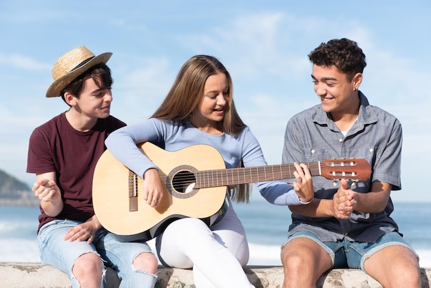 Grupa szczęśliwych, wieloetnicznych nastolatków śpiewających i grających na gitarze siedzących nad morzem