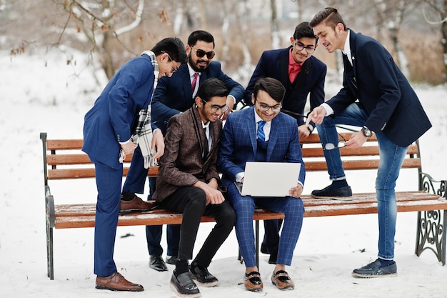Grupa sześciu indyjskich biznesmenów w garniturach pozowanych na zewnątrz w zimowy dzień w Europie patrząc na laptopa