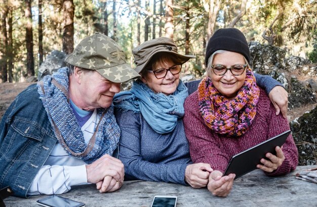 Grupa Uśmiechniętych Seniorów Patrzących Na Cyfrowy Tablet Siedząc Przy Drewnianym Stole W Górskim Obszarze Piknikowym, Ciesząc Się Plenerem, Spędzając Razem Czas Koncepcja Technologii I Komunikacji