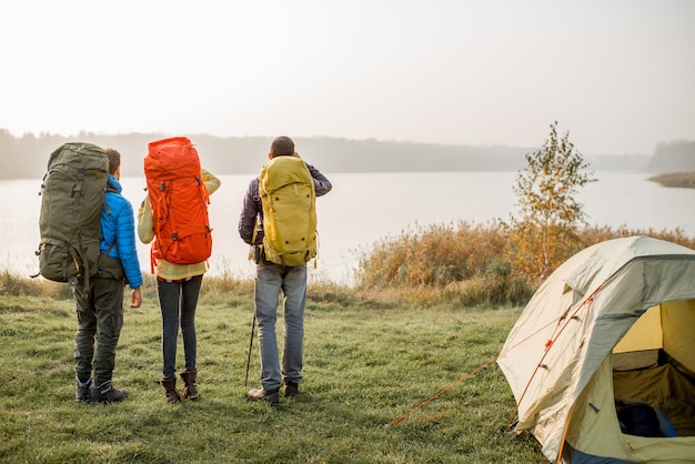 Grupa wędrowców z kolorowymi plecakami stojąca z tyłu nad jeziorem w pobliżu kempingu podczas mglistej pogody
